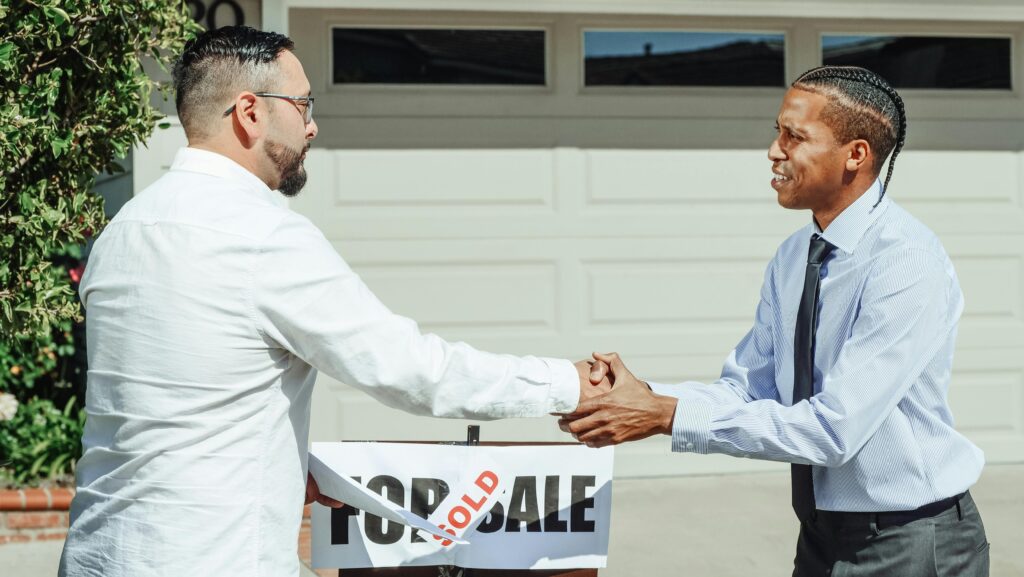 Two men shaking hands in front of a sold house sign, sealing a real estate transaction.